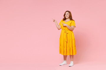 Full length portrait smiling young redhead plus size body positive female woman 20s in yellow dress pointing index fingers aside on mock up copy space isolated on pastel pink color background studio.