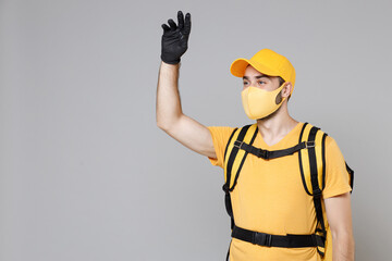 Delivery employee man guy in yellow cap t-shirt uniform face mask gloves thermal food bag backpack work courier service during quarantine coronavirus covid-19 virus isolated on gray background studio.