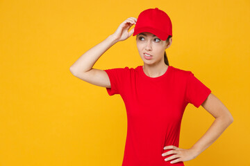 Delivery employee woman in red cap blank t-shirt uniform workwear work courier in service during quarantine coronavirus covid-19 virus confused look aside isolated on yellow background studio portrait