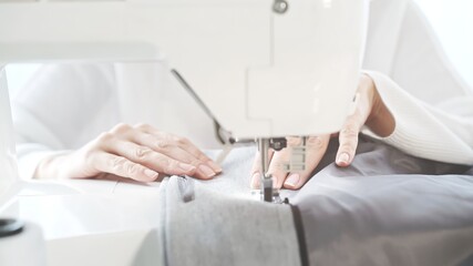 Female hands on a white sewing machine close-up. Concept of sewing in modern bright studio, woman in white sweater sewing grey cloth in process