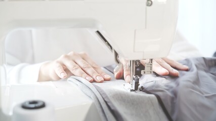 Female hands sew on a white sewing machine close-up. Concept of sewing in modern bright studio, woman in white sweater sewing grey cloth in process, spool of thread on foreground