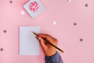 Golden Holiday decorations and notebook in woman's hands on pink background, flat lay style. Christmas planning concept.