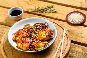plate of soba with chicken and sesame seeds in teriyaki sauce on a wooden stand and sticks next to sesame seeds and soy sauce.