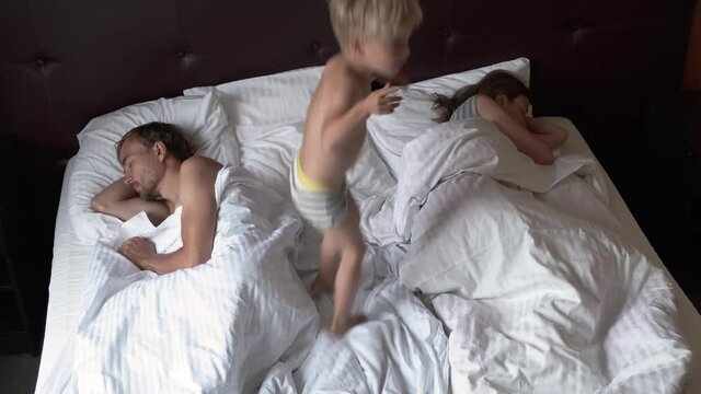 Funny annoying little child boy climbs on bed and start to jumping to wakes up sleepy parents. Morning routine in happy loving family.