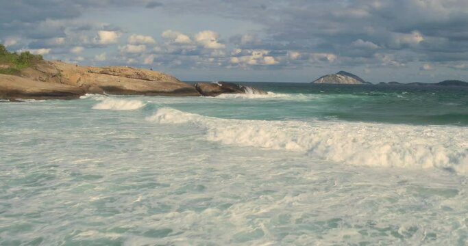 Low altitude flyover of frothy waves on sunny day & wispy sky, Arpoador beach