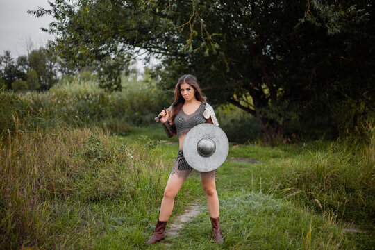 A medieval female warrior dressed in chain mail with a sword and shield in her hands poses against the background of a forest. Fantasy costume, combat makeup.