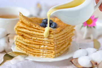 A stack of pancakes with blueberries poured with condensed milk on the background of a cup of tea, a tablecloth, cotton and a gold fork