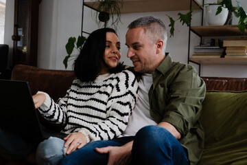 Fototapeta premium Romantic couple smiling at each other while waiting to chat with friends on video call using laptop sitting on sofa in lounge.
