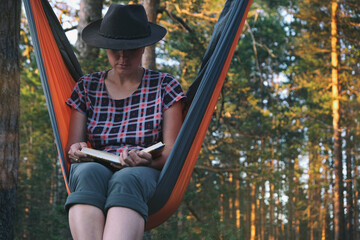 Woman in fedora hat reading book while sitting in hammock in pine forest at sunset. Mindfulness, relax on nature, treat yourself, digital detox, unplugging, life offline concept.