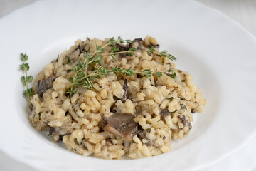 Closeup view of creamy risotto or Italian arborio rice dish with mushrooms, thyme, white wine and parmesan cheese served in plate with fork on white wooden background. Image with selective focus