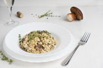 Creamy risotto or Italian arborio rice dish with broth, mushrooms, thyme, olive oil and parmesan cheese served in plate with glass of white wine on white wooden background. Image with copy space