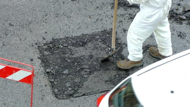 Workman in white coverall uniform installing cement solution on destroyed part of pavement road using iron shovel. Process of casting cement on construction site, engineering concept