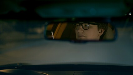 Driver's face in eyeglasses is reflected in a rear view mirror, night shot. Driver in a mirror in car, night road shot driving all night from backseat
