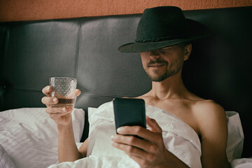 Brutal unshaven man in cowboy fedora hat is lying in bed, drinking whiskey from glass and reading...