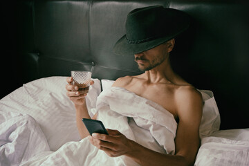 Brutal unshaven man in cowboy fedora hat is lying in bed, drinking whiskey from glass and reading messages on smartphone. Alcoholism, hangover or sad birthday party alone. Dating online concept.