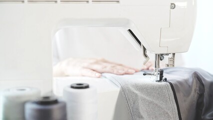 Female hands sew on a white sewing machine close-up. Concept of sewing in modern bright studio, woman in white sweater sewing grey cloth in process, spool of thread on foreground