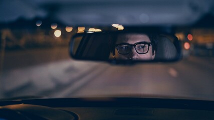 Driver's face is reflected in a rear view mirror, night shot, violet color, bokeh light. Tired concentrated driver in eyeglasses is reflected in a mirror in car, night road shot driving all night