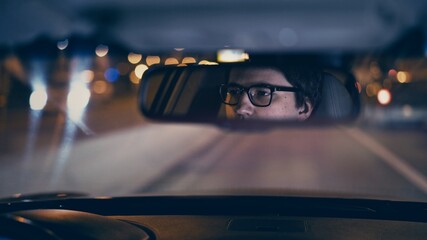 Driver's face is reflected in a rear view mirror, night shot, violet color, bokeh light. Tired concentrated driver in eyeglasses is reflected in a mirror in car, night road shot driving all night © diproduction