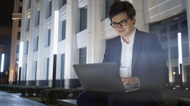 Man With Laptop On Lap Smiling Sitting On Bench Outside The University. Learning Outside At Night Time, Man In Black Jacket Alone Typing In Laptop, Middle Shot