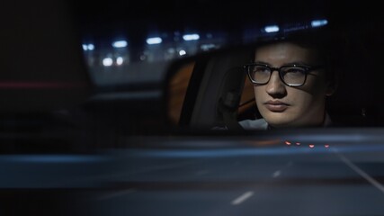 Driver's face is reflected in a rear view mirror, night shot, dark blue road. Driver in eyeglasses...