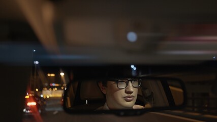 Driver's face is reflected in a rear view mirror, night shot. Driver wearing eyeglasses is reflected in a mirror in car, night road shot driving all night