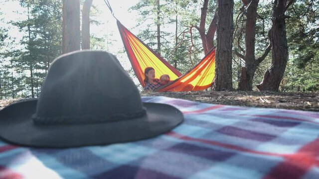 Mother and child resting in tourist hammock in pine forest, fedora hat lying on picnic plaid on foreground. Camping, hiking, active family, outdoor gear.