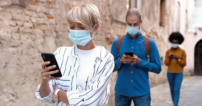 Portrait of multi-ethnic young people standing in queue and tapping on mobile phones. Beautiful Caucasian woman and man in medical masks texting on smartphones. African American woman in quarantine