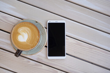 Large mug with aromatic cappuccino coffee and white smartphone on a wooden table