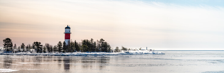 Panorama, close view of red white lighthouse in middle of frozen, snowed island at cold Baltic Sea, partly open water, thin ice reflecting daylight. Blue sky, light breeze. Northern Sweden, Umea