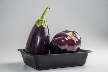 Two oblong and round eggplants in a black plastic rectangular plate on a solid background.