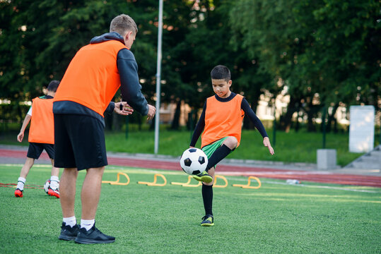 Motivated kid player in football uniform working out the kicking ball together with his experienced coach on sport field