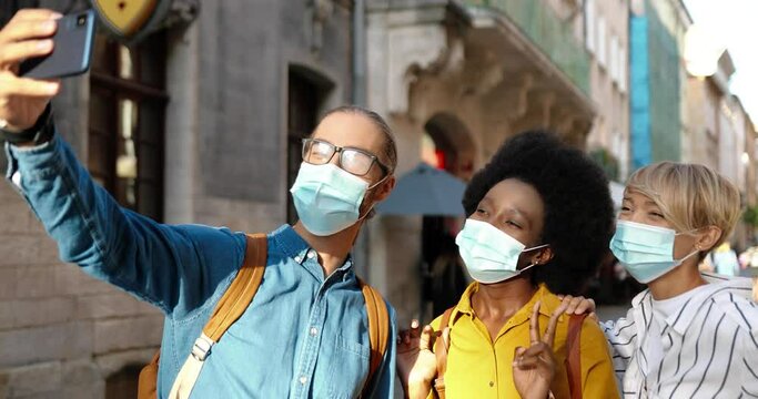 Close up portrait of mixed-races friends in medical masks standing in city and making selfie photos on smartphone. Joyful Caucasian guy making video on street with woman and African American woman