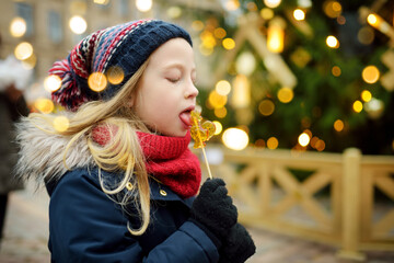 Cute young girl having rooster-shaped lollipop on traditional Christmas fair in Riga, Latvia. Child enjoying sweets, candies and gingerbread on Xmas market.