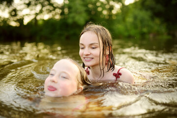 Two young sisters having fun on a sandy lake beach on warm and sunny summer day. Kids playing by the river.