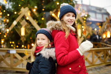 Two adorable sisters having rooster-shaped lollipops on traditional Christmas fair in Riga, Latvia. Children enjoying sweets, candies and gingerbread on Xmas market.