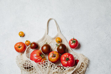 Fresh colorful heirloom veriety tomatoes in eco friendly mesh shopping bag on white concrete background. Zero waste