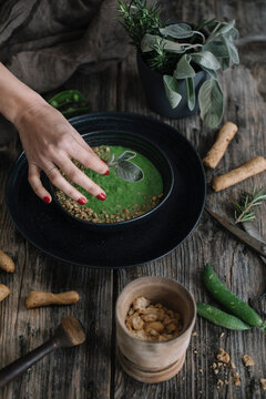 Hand From Above Garnishing Green Peas And Herb Soup With Breadsticks