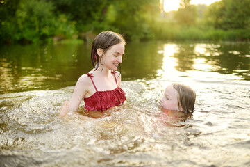 Two young sisters having fun on a sandy lake beach on warm and sunny summer day. Kids playing by the river.