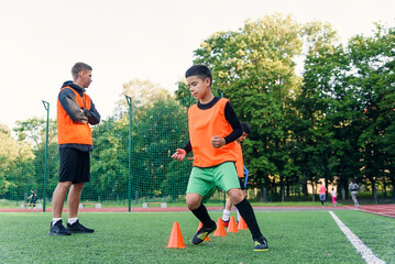 Concentrated junior soccer player running among orange cones that standing on artificial turf at...