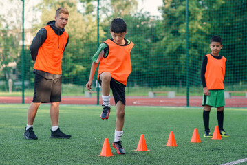 Concentrated junior soccer player running among orange cones that standing on artificial turf at...