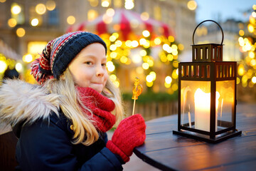 Cute young girl having rooster-shaped lollipop on traditional Christmas fair in Riga, Latvia. Child enjoying sweets, candies and gingerbread on Xmas market.