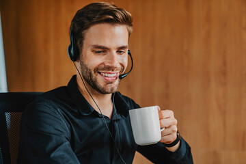 Young man in headset holding coffee cup working in office, coffee break hour concept