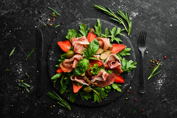Salad of arugula, prosciutto, strawberries and capers on a black stone plate. Italian cuisine.