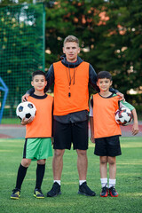 Happy teen football players standing on the green soccer field with the coach and looking at camera with smiles. © gorynvd