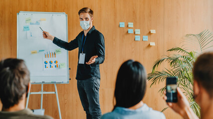 Young adult businessman with protective face mask talking about new project while giving presentation to group of colleagues in board room.