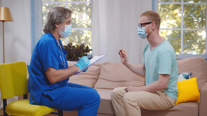 Senior doctor in protective mask writing prescription in checklist document talking with patient
