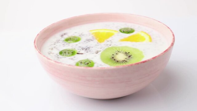 Rotating of yogurt with kiwi, gooseberry, chia in ceramic bowl on white background. Side view, close up.