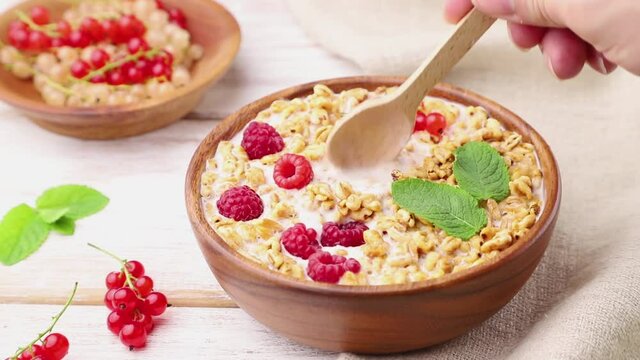 Wheat flakes porridge with milk, raspberry and currant in wooden bowl on white wooden background and linen textile stir with a spoon. Side view, close up.
