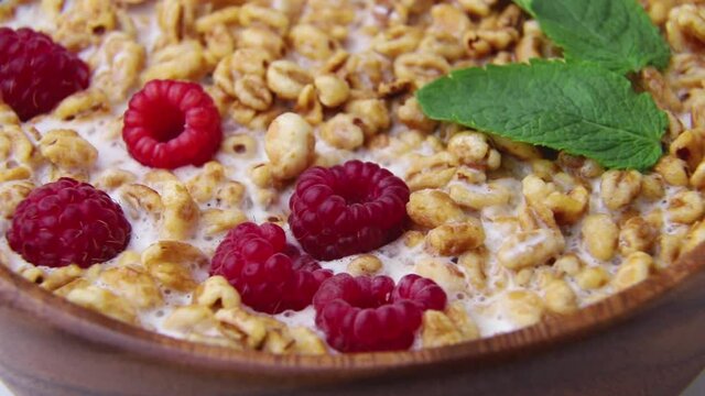 Rotating of wheat flakes porridge with milk, raspberry and currant in wooden bowl on white wooden background and linen textile. Side view, close up.