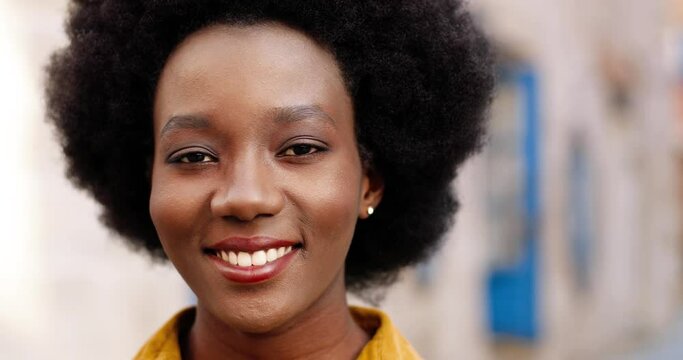 Close up portrait of cheerful African American beautiful young female smiling to camera while standing outdoors. Pretty joyful girl with smile on face posing in town. Positive emotions concept